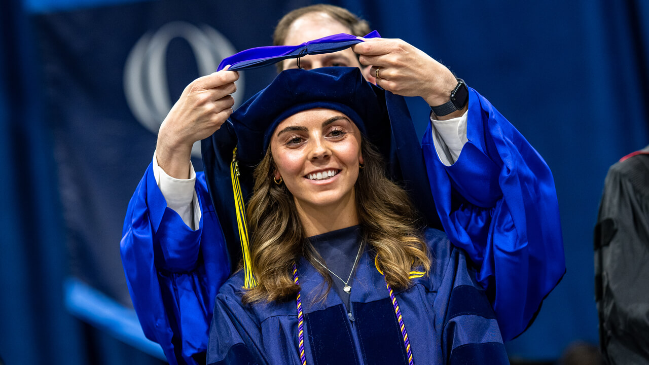 A graduate smiles as a faculty member lifts a purple hood over her head