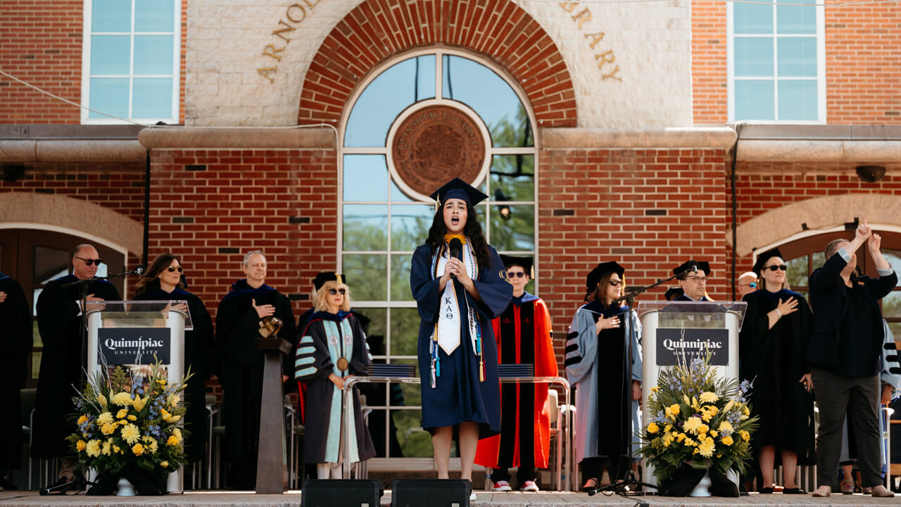 Gabriela Merino sings with a microphone on the library steps with administrators behind her
