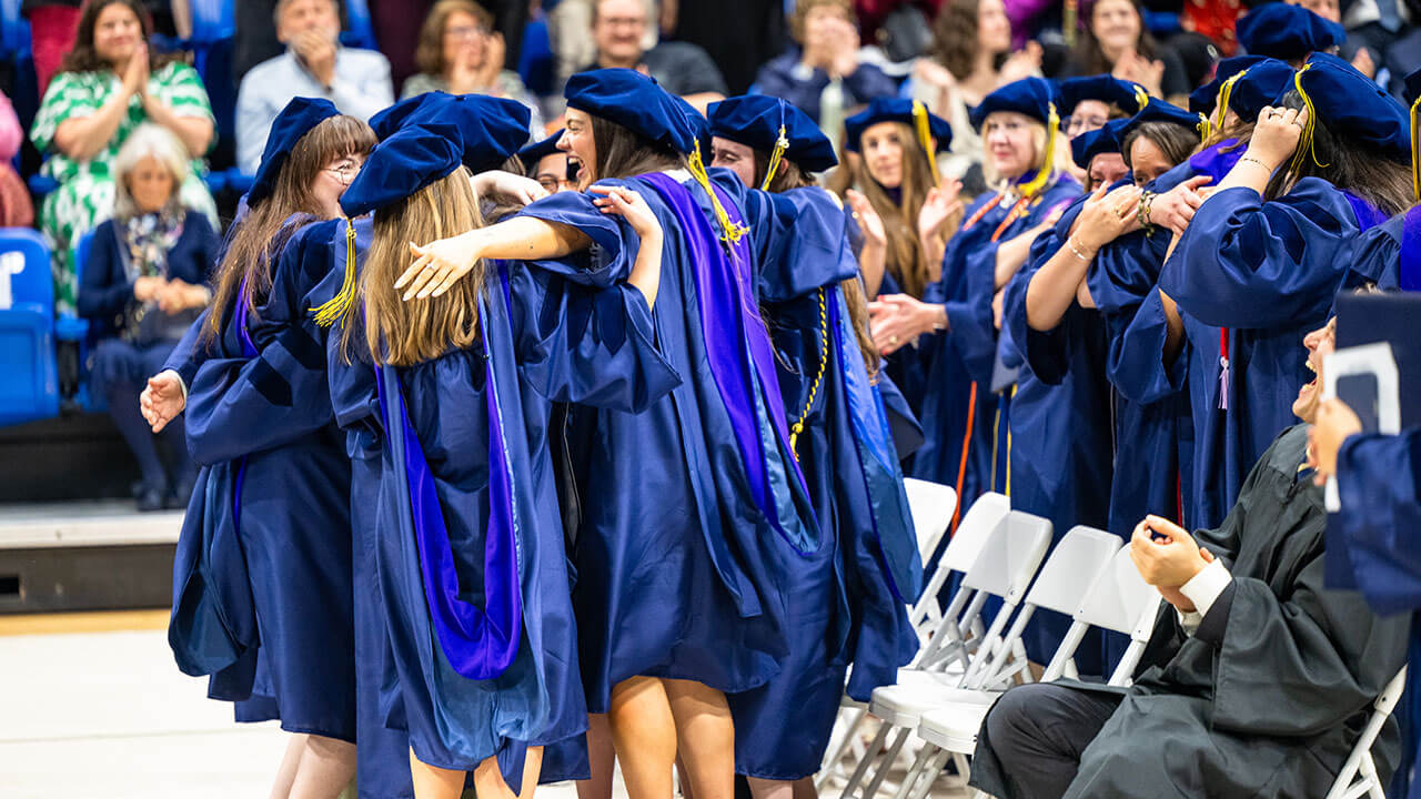 A group of graduates dressed in regalia hug during commencement