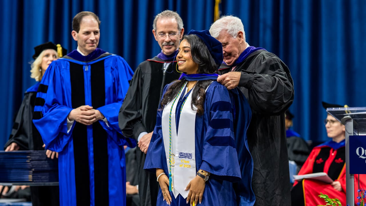 A faculty member places a hood over the neck of a graduate who smiles on stage