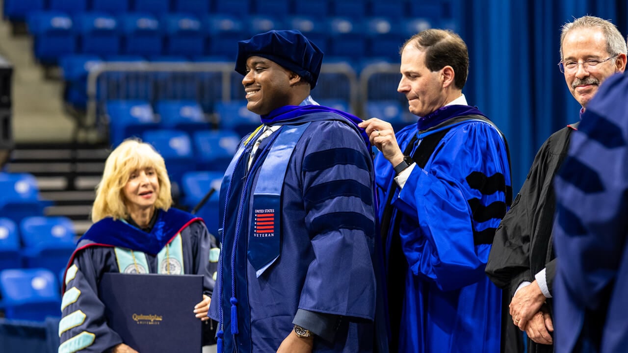 A graduate wearing a U.S. Veteran stole smiles as he receives his doctoral hood from a professor