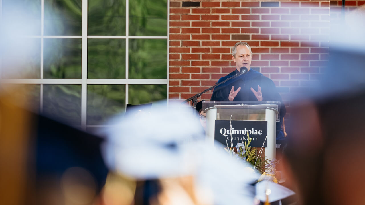Graduates sit and listen to Richard Benton as he speaks at a podium on the library steps
