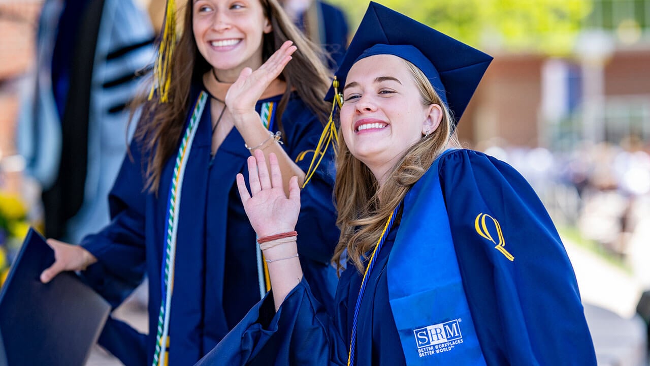Two students smiling and waving in their graduation regalia