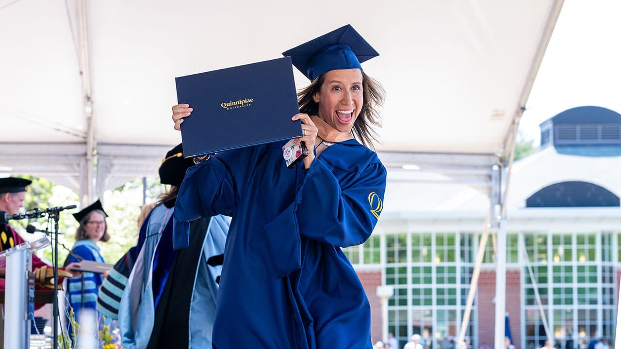 A graduate on commencement stage holding up their diploma in excitement