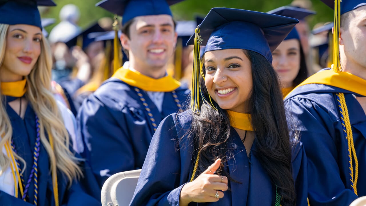 A graduate smiles and gives a thumbs up as she sits in a row of graduates