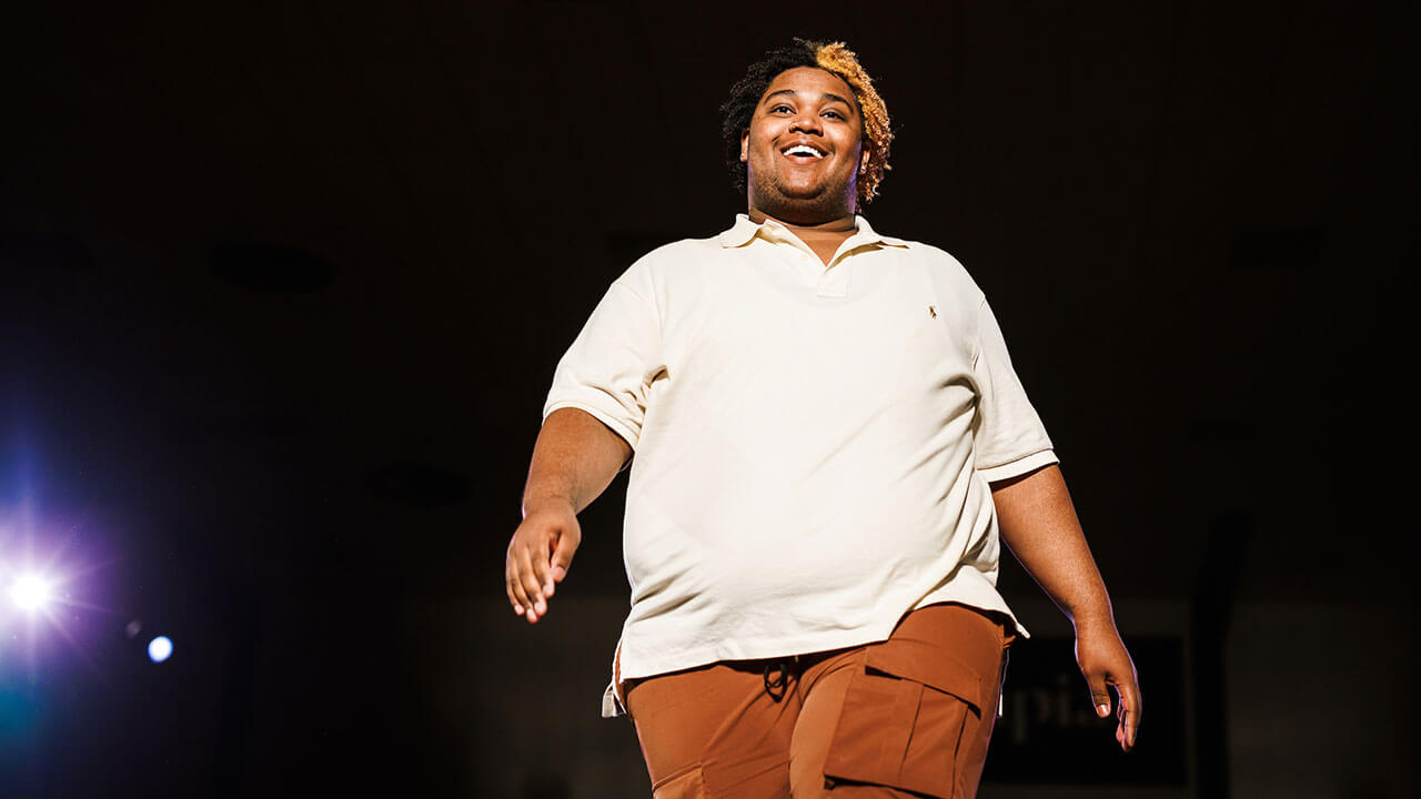 black man with his hair half black and half blonde walks down the runway in a white shirt and brown pants