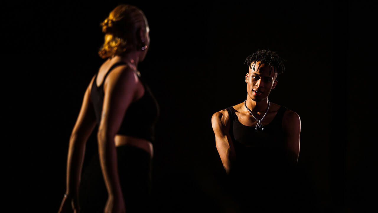 black man faces the camera wearing all black while a black woman faces him, also in all black