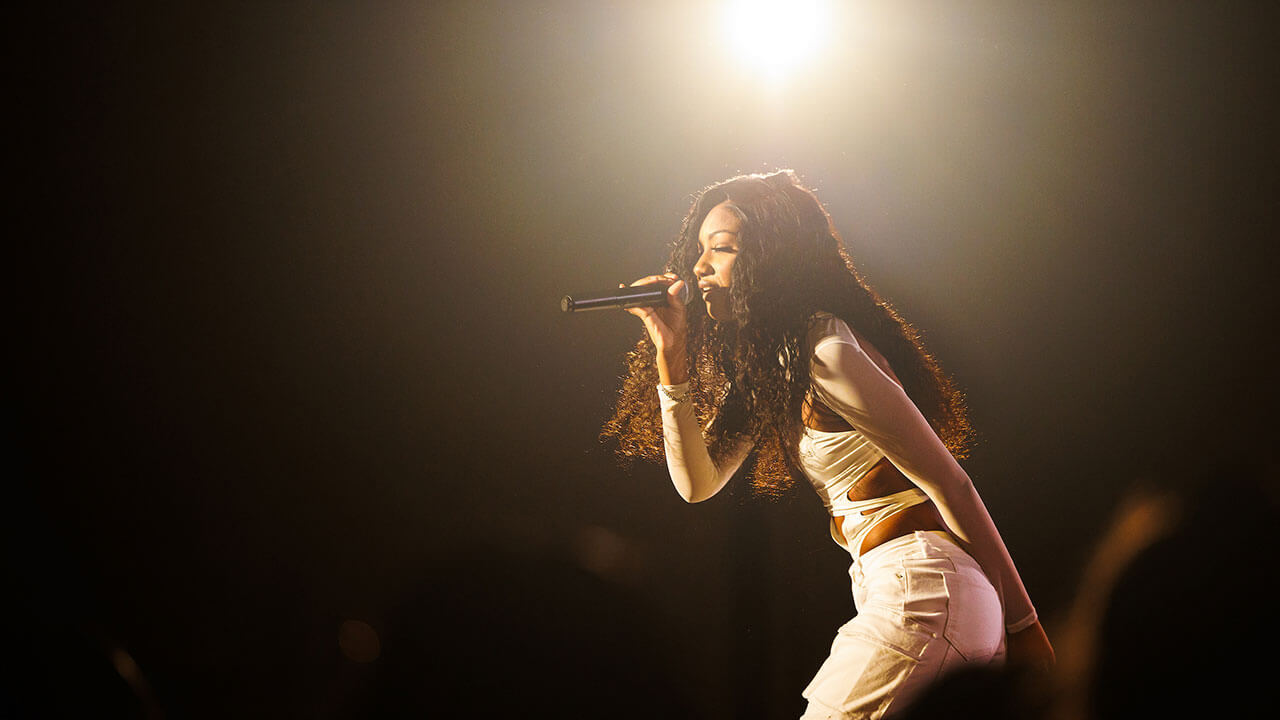 black singer in a white dress with long braids leans forward in front of a spotlight