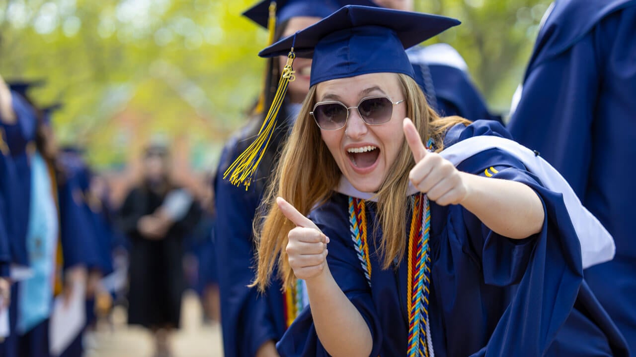 Graduate smiles with two thumbs up as they process into commencement.