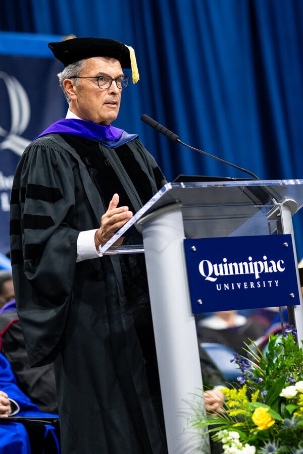 Craig McLean in a doctoral cap and gown speaks at a Quinnipiac podium with flowers in front of it on stage