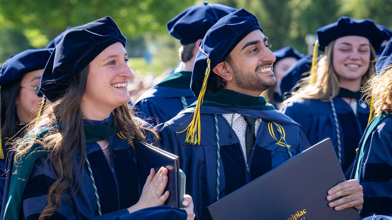 Graduates stand, clapping and smiling with their diplomas.