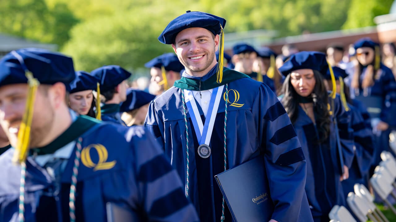A graduate with a doctoral cap and wearing a Quinnipiac medal on his neck smiles