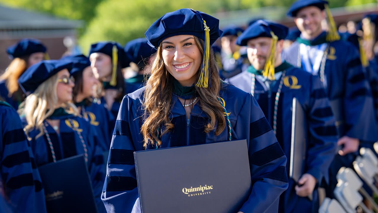 A graduate in a doctoral cap holds her diploma and smiles broadly
