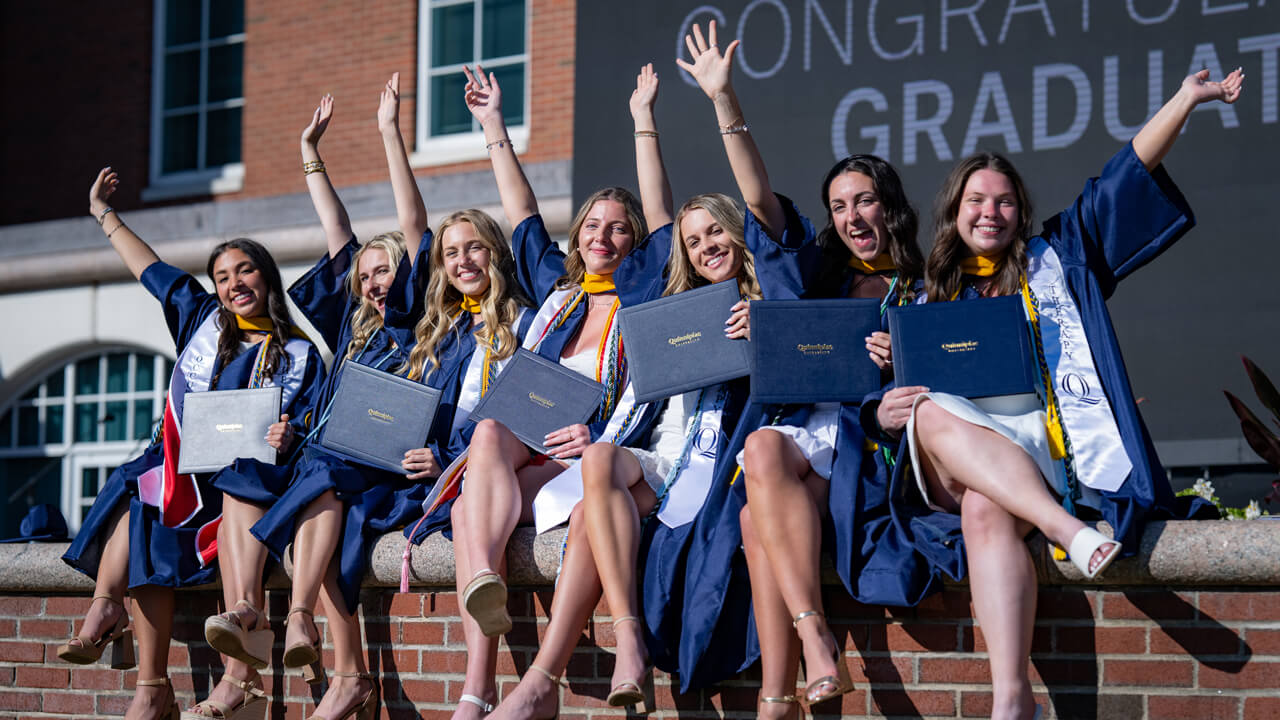 7 graduates sit along the library wall and hold their arms and diplomas up in celebration