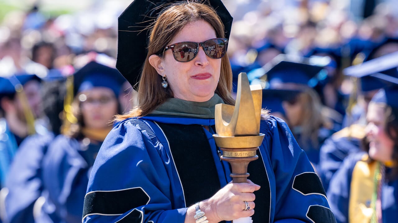 Karen Majeski wears doctoral robe and cap and carries the Commencement mace down the path