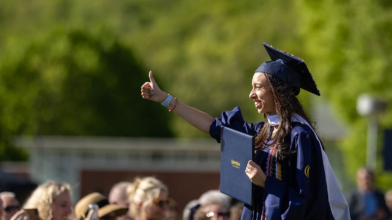 A graduate holding their diploma and holding their thumb up