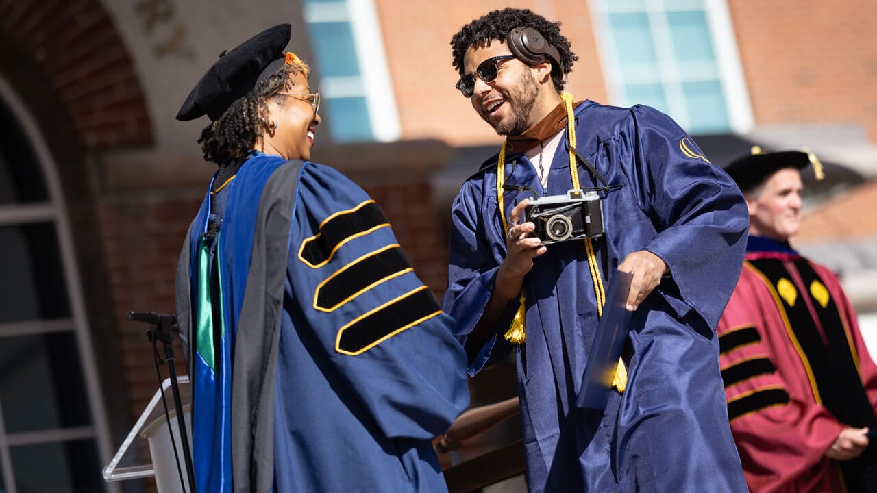 Graduate accepts diploma with a camera in their hand.