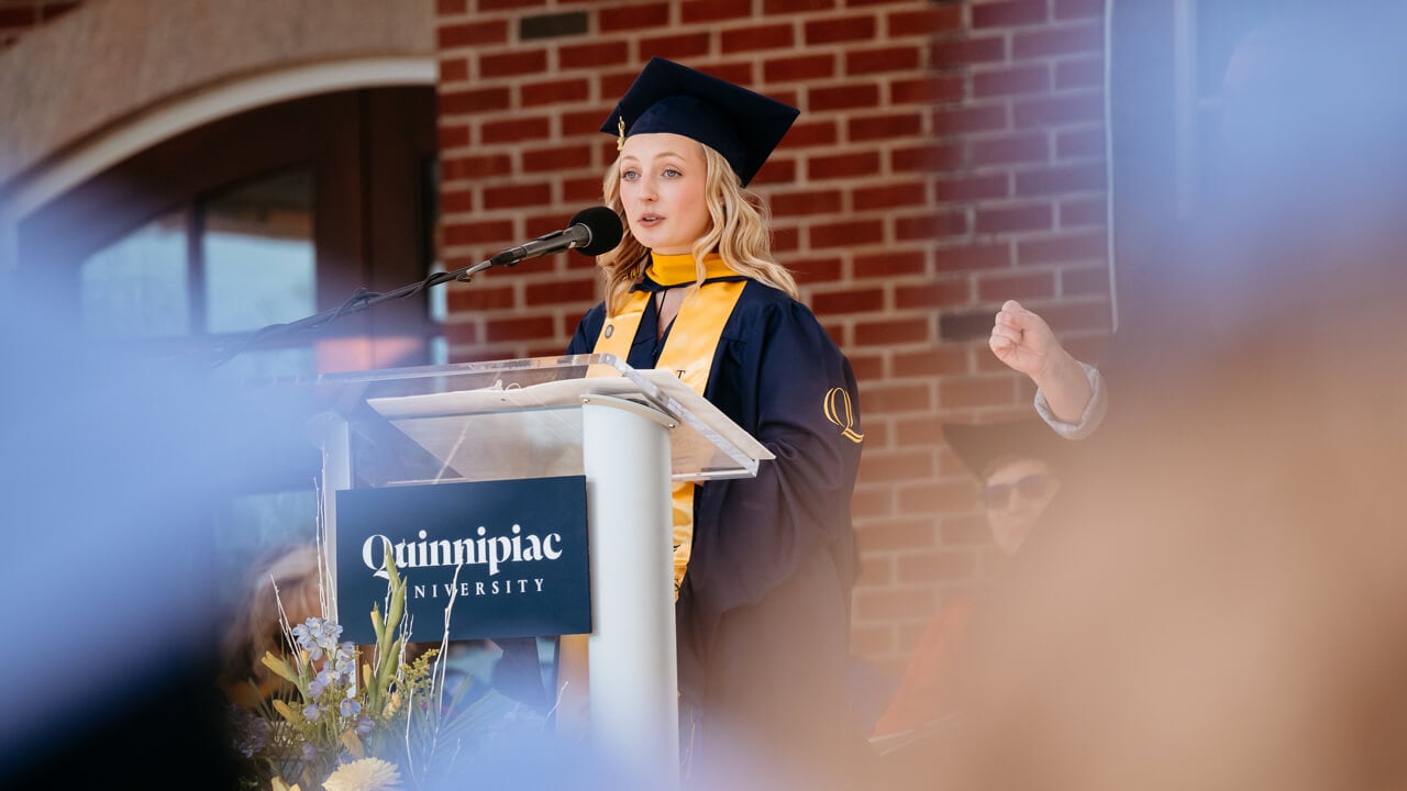 Kathryn Damato in her cap and gown speaks at a Quinnipiac podium on the library steps
