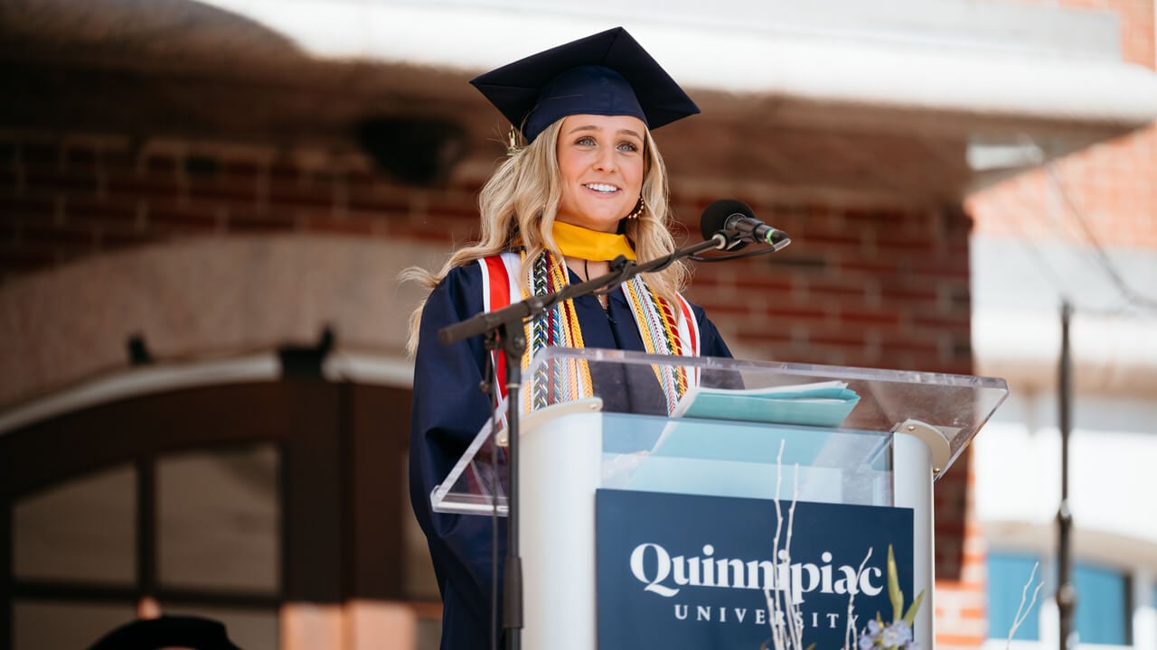 Olivia Ferreira wears a cap, gown and many cords as she speaks at the podium