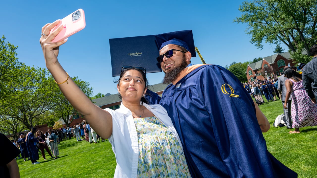 A graduate holds up a diploma and poses for a selfie with a loved one