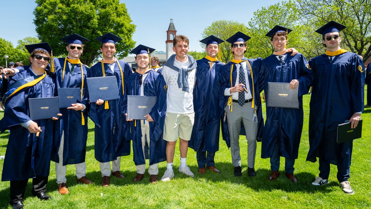 8 graduates pose arm in arm for a photo in front of the library clocktower
