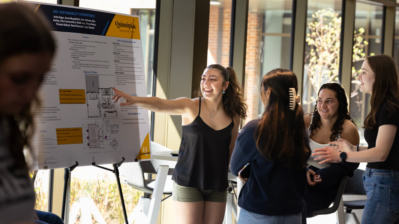 A student in The Grove points to a poster presentation board while other students look on
