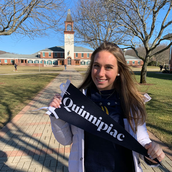 Photo of a prospective student holding a Quinnipiac flag on the quad with the Arnold Bernhard Library in the background