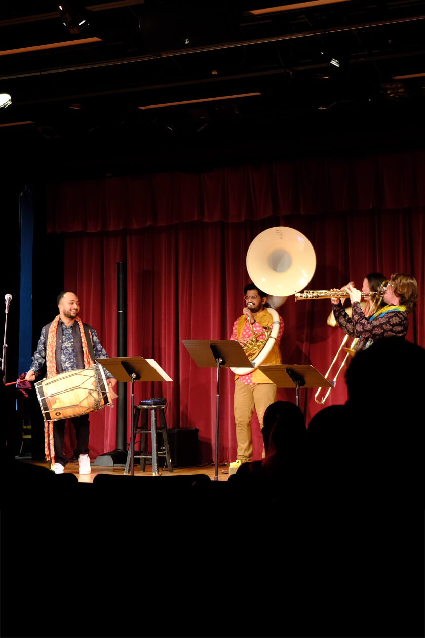 Butter Chicken performs live on stage with a sousaphone, dhol drum, and brass instruments against a red curtain backdrop.