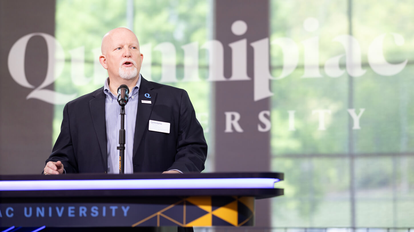 John A Bau, Jr. Assistant Dean for Student Experience and Career Development  addresses the audience at a podium bearing the Quinnipiac University logo.