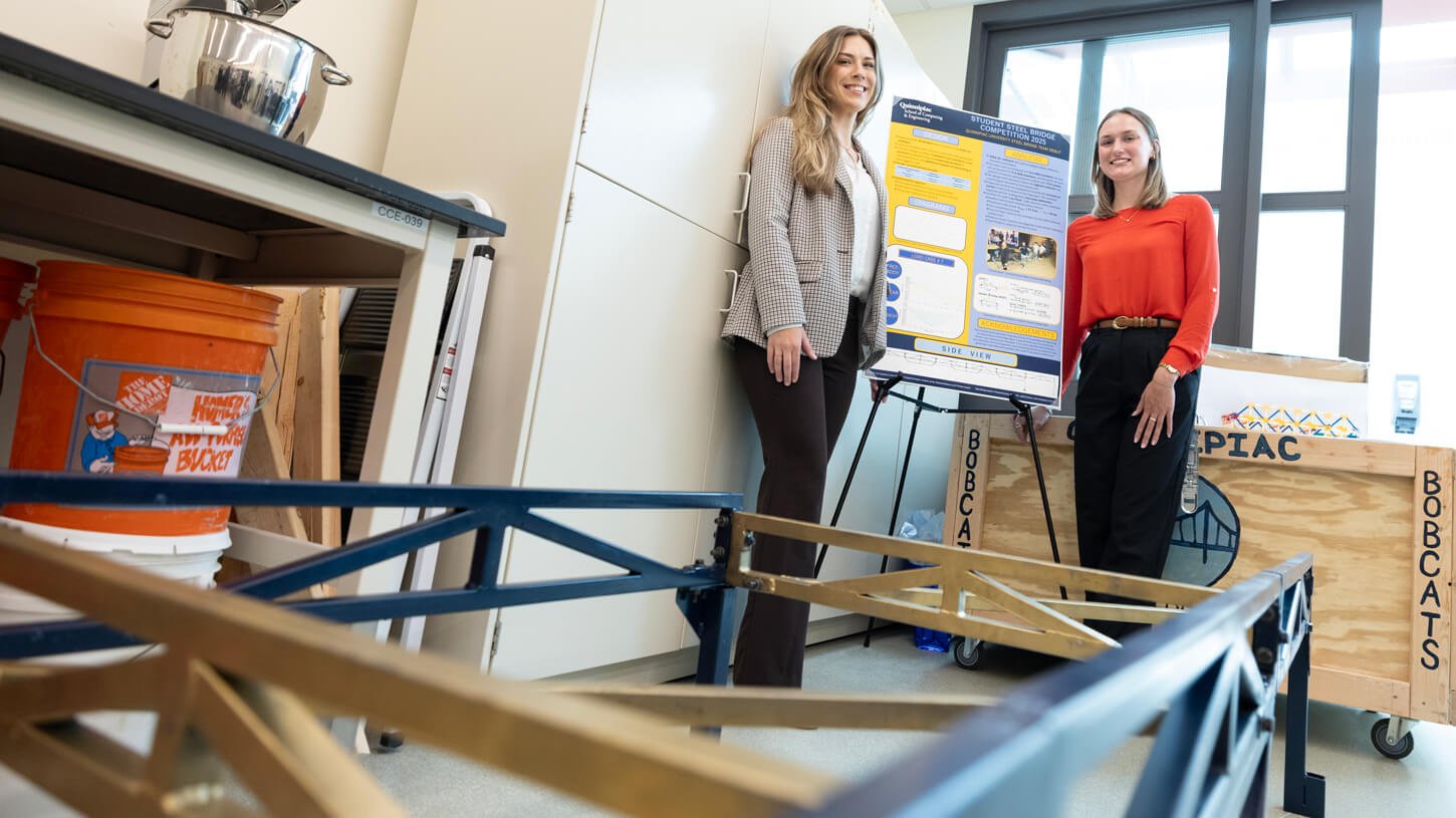 Two students stand beside their bridge-building project and informational poster inside an engineering lab.