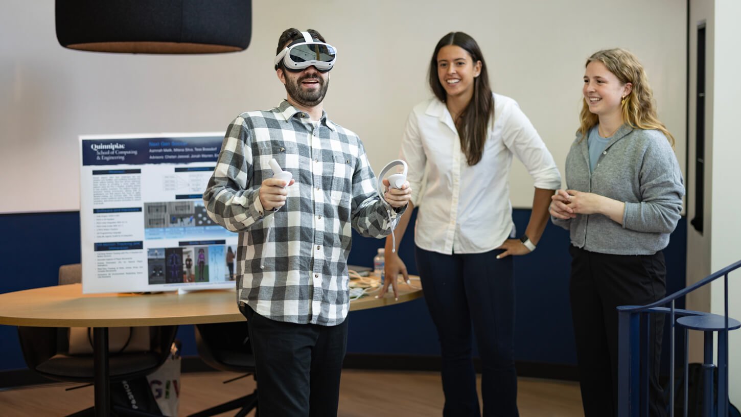 A professor wearing a VR headset and holding controllers plays a demo of a virtual reality project while two classmates stand nearby smiling.