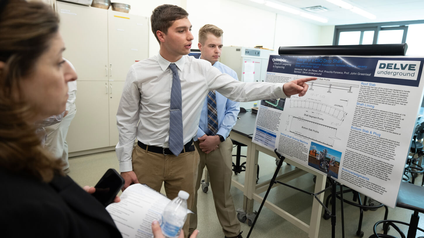 A student points to a technical diagram on a poster explaining a drop shaft design project, while another student and a visitor observe.