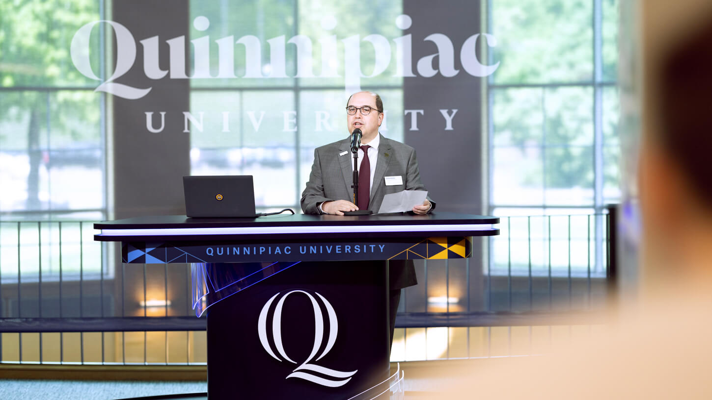 Taskin Kocak, PhD, speaks at the open-air studio podium branded with the Quinnipiac University logo during the Projects Day event.