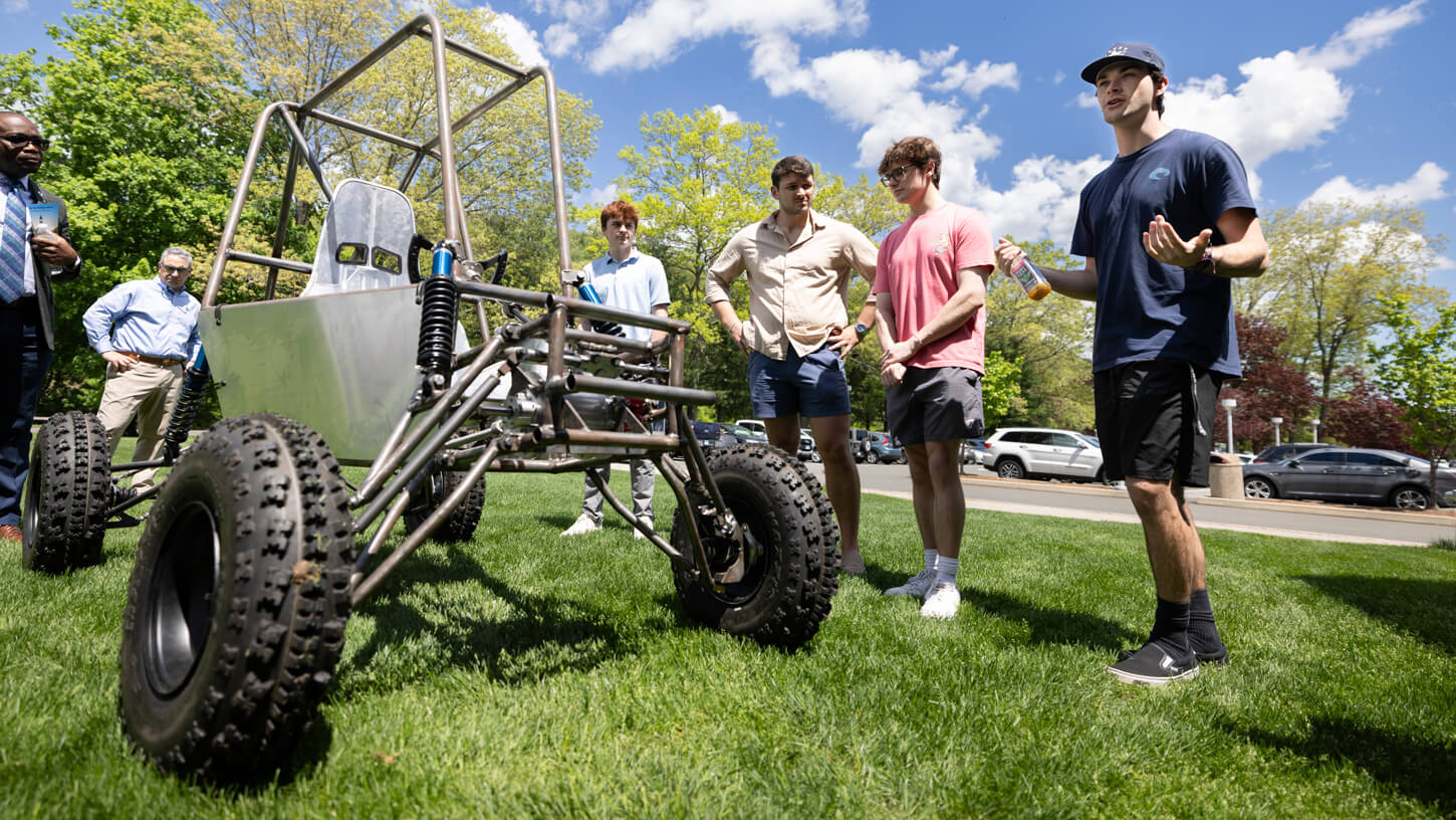 A team of students presents an off-road baja vehicle prototype on the campus lawn to faculty and visitors.