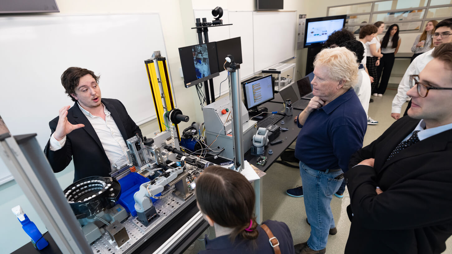 A student presents a robotics project involving a complex mechanical setup to a group of onlookers in a lab setting.