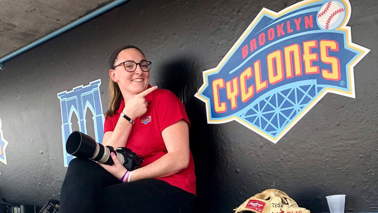 Photographer smiles with a camera next to a sign that reads Brooklyn Cyclones.