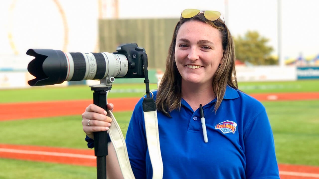 Photographer smiles with a camera on a baseball field.