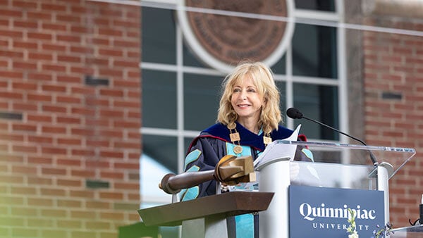 President Judy Olian dressed in academic regalia stands on stage at the podium at a Quinnipiac commencement ceremony.