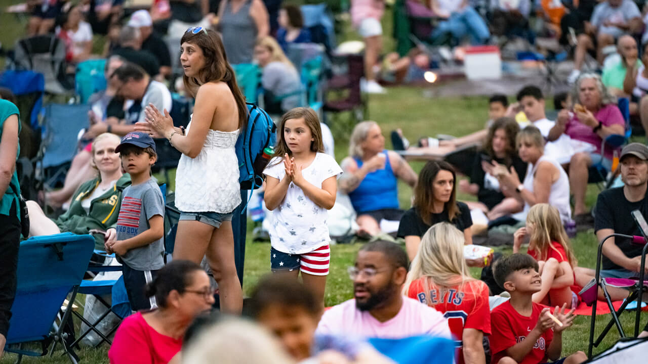 Dozens of family members in patriotic outfits clap from the grass on the Hamden town green