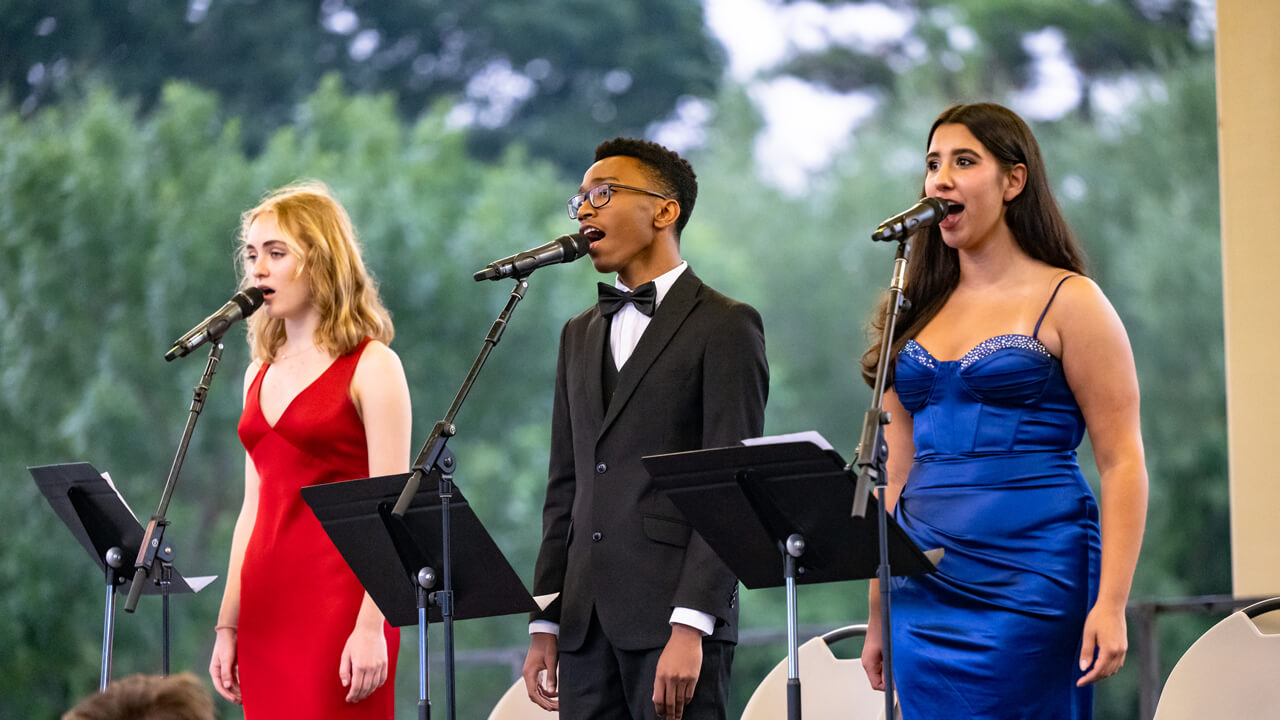 Three singers, one in red, one in black and white and one in blue sing at microphones