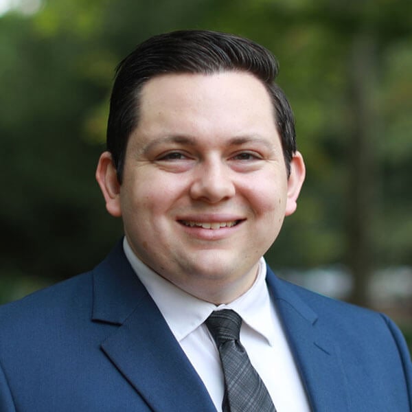 John Martin smiles in a navy suit against some greenery