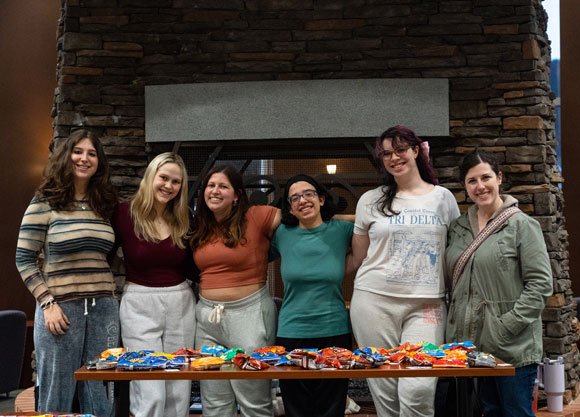 Students stand behind a table and pose for a photo in the Piazza.