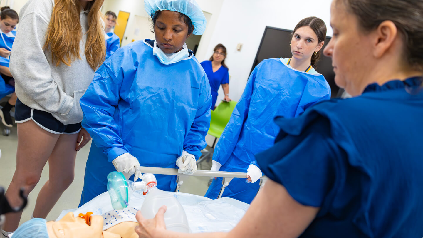 Students gather around a medical mannequin as an instructor demonstrates how to use an oxygen bag valve mask during a hands-on lesson in a clinical lab.