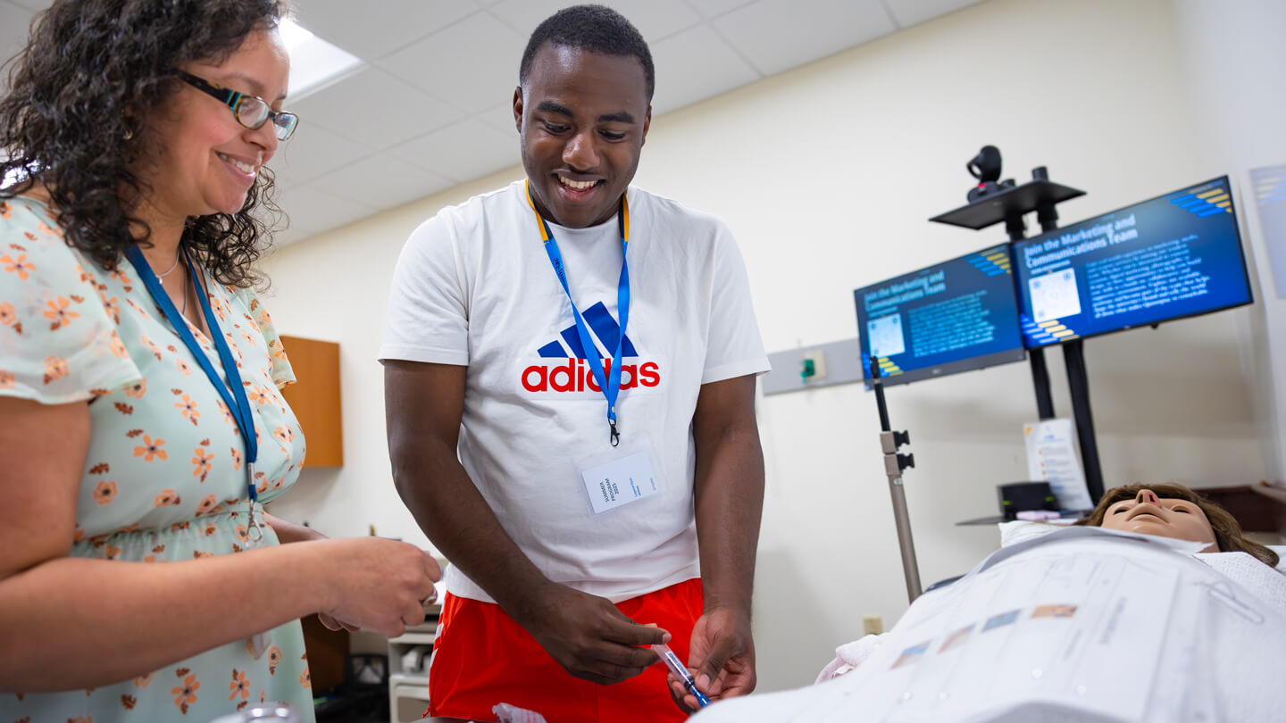 A student smiles while learning to administer an injection to a medical mannequin, guided by an instructor during a simulation activity.