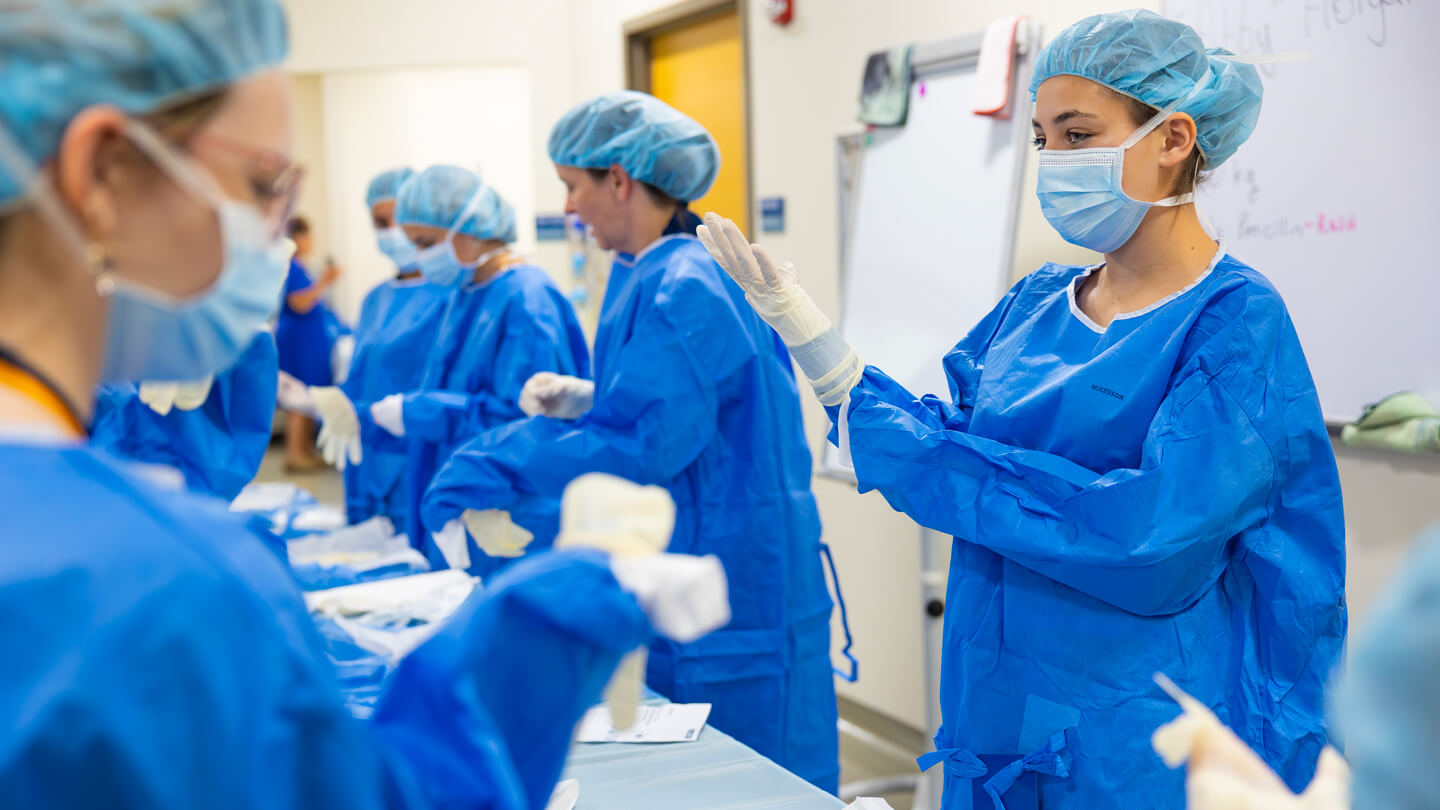 High school students in blue surgical gowns and masks practice sterile glove techniques during a nursing summer camp session at Quinnipiac University.