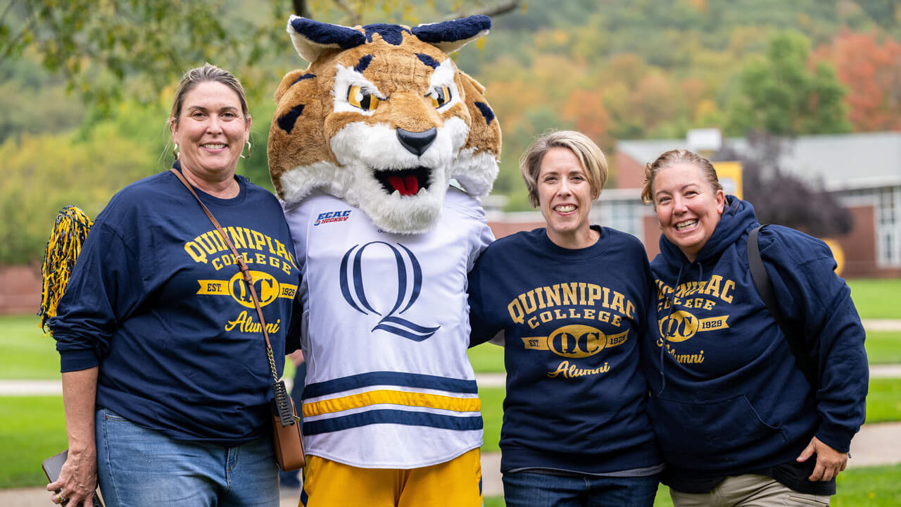 Three Quinnipiac alumni in matching shirts stand together with the mascot Boomer the Bobcat.
