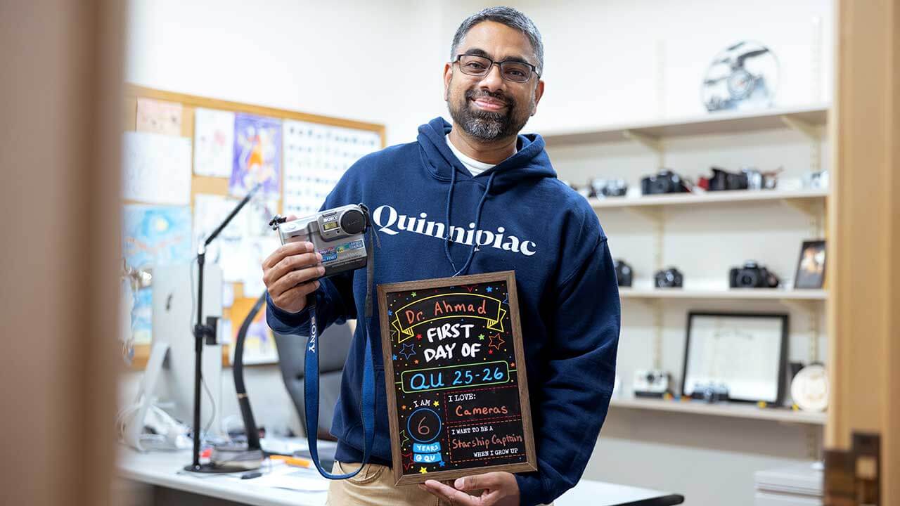 Wasim Ahmad holds up a camera alongside a first day of classes sign
