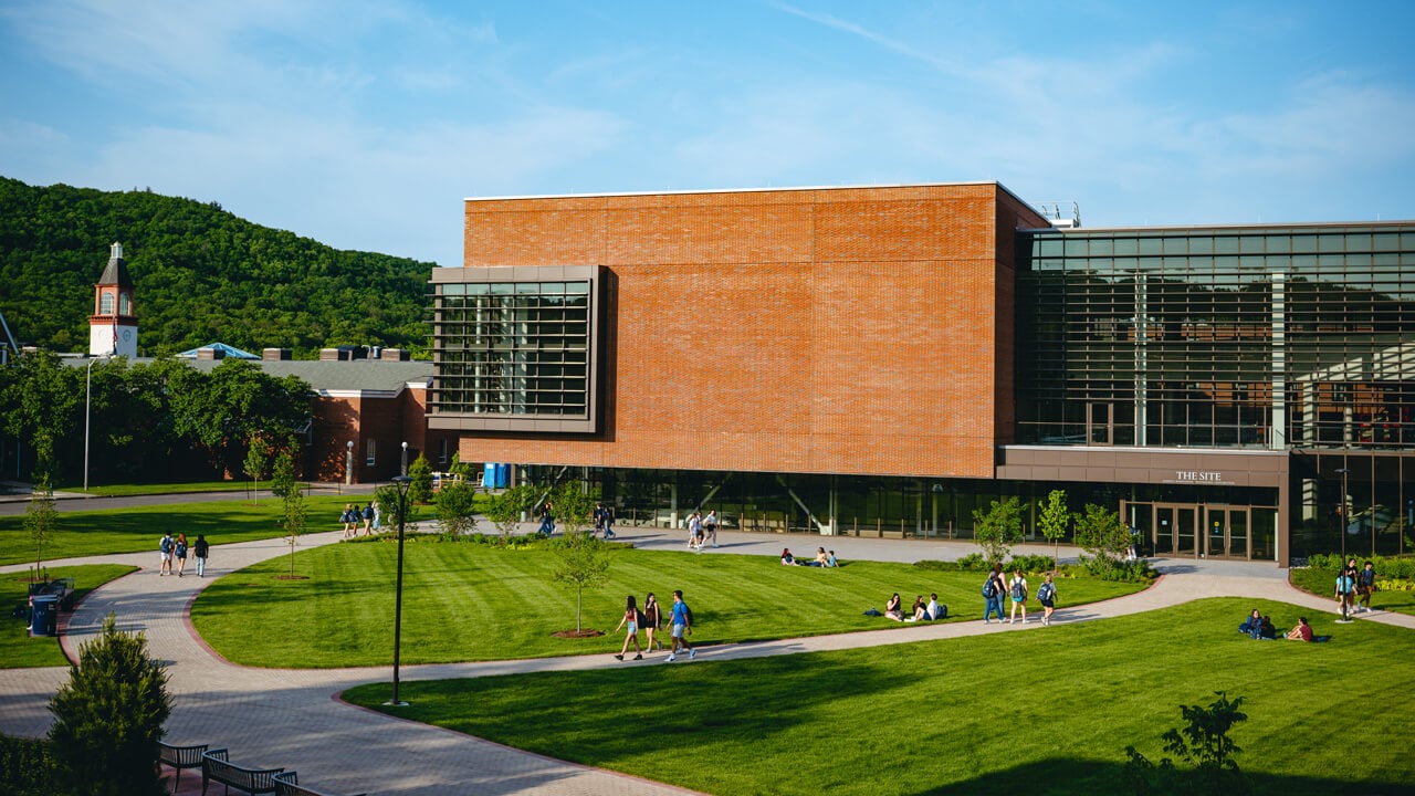 Students walk on paths and sit on grass in front of The SITE with the library clocktower in the background