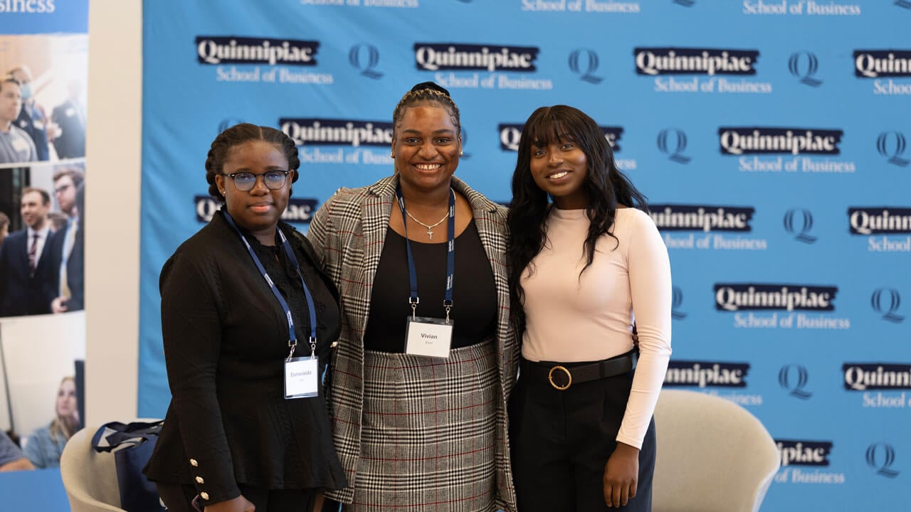 Students smile against a blue Quinnipiac step and repeat