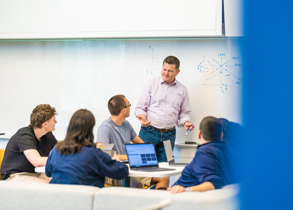 a male professor talking to a group of 4 graduate students sitting at a table in a classroom with writing on the whiteboard